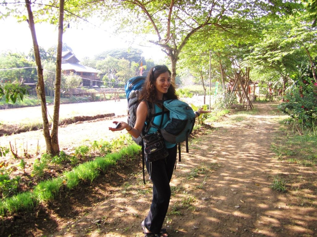 Kate at age 26 in 2010, wearing a large backpack on her back and a small backpack in front, double turtle style, standing on a wooded path in Pai, Thailand.