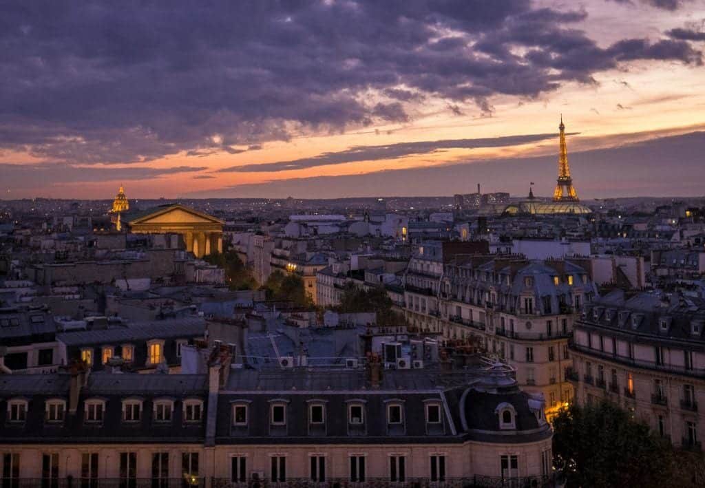 A purple cloudy sunset in Paris. You see the rooftops in the fading light and on the right, in the distance, the Eiffel Tower lit up in orange.