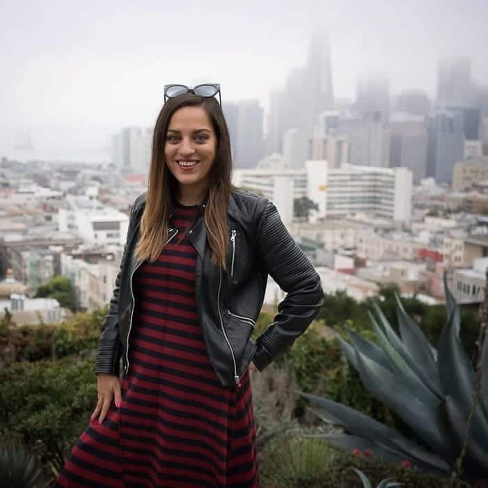 Kate wearing a burgundy and navy striped long dress and short black leather jacket in front of the San Francisco skyline, covered by fog.