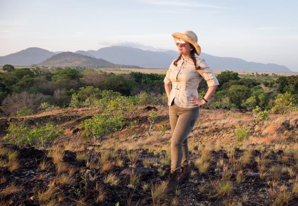 Kate wearing a tan jacket and green pants and boots, and a hat, looking out over the savannah grasslands in the Rupununi of Guyana.