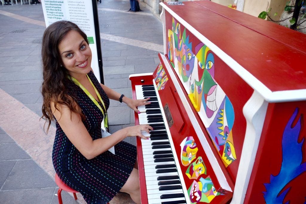 Kate is sitting down and playing a bright red painted piano in the streets of Trento. She looks at the camera and smiles.