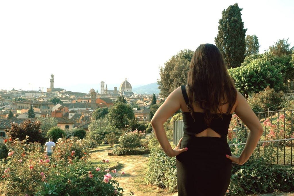 Kate faces away from the camera and stands facing the Duomo in Florence in the distance. She is in a rose garden, surrounded by greenery.