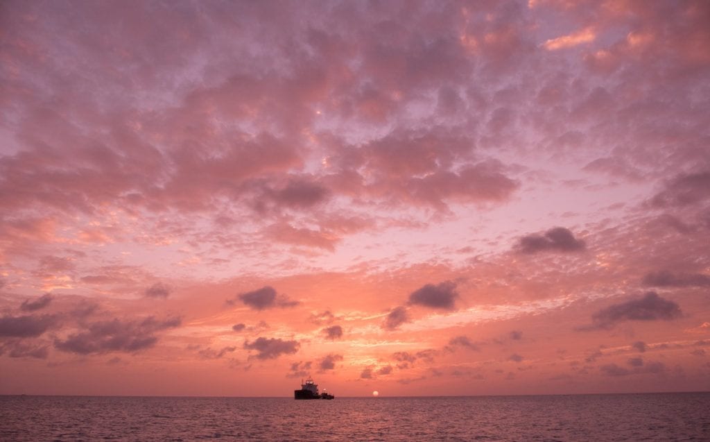 A pink sunset with lots of fluffy purple clouds in Antigua. At the bottom is the sea, a black silhouette of a boat, and a tiny yellow sun.