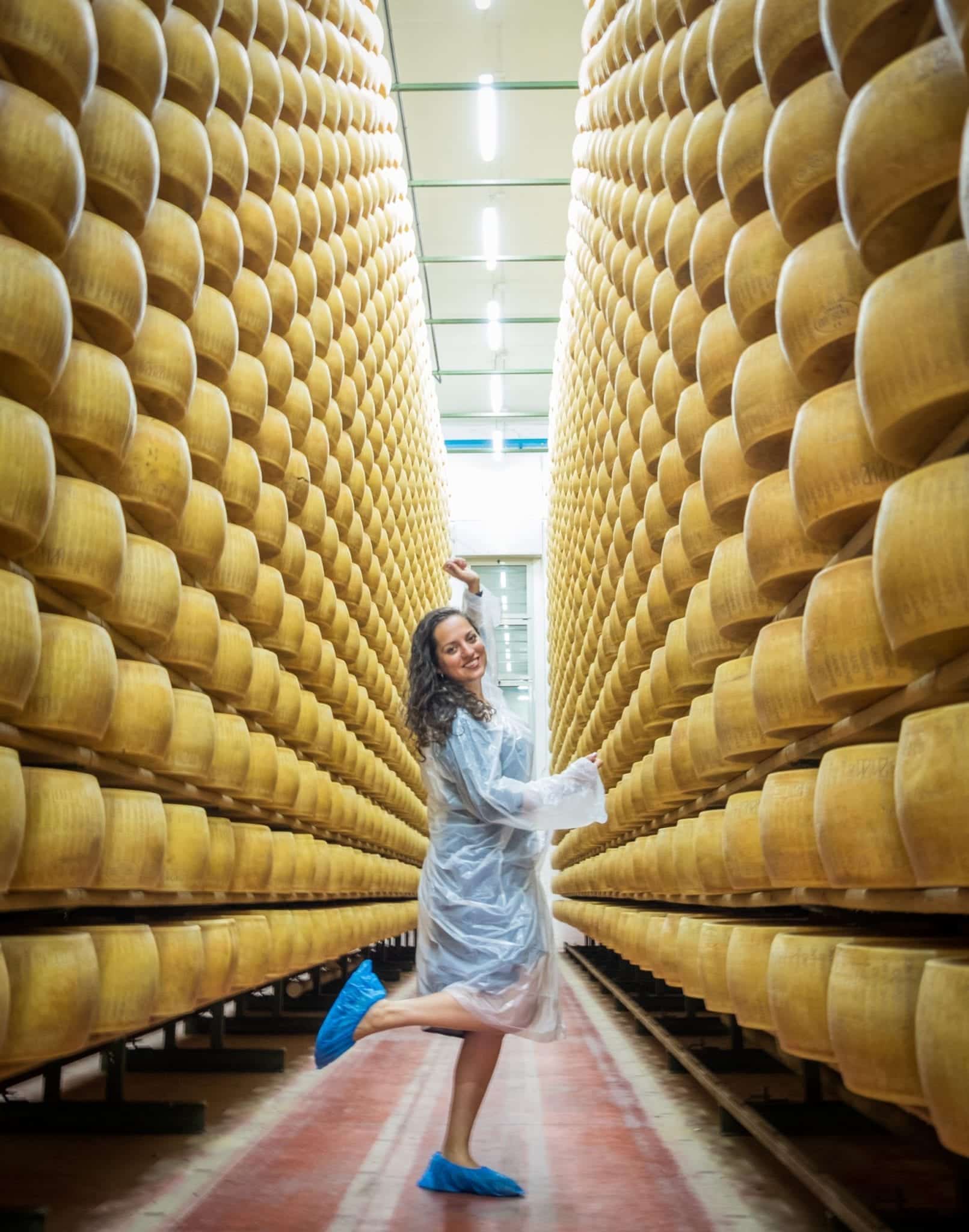 Kate poses with one foot in the air in the middle of the aisle in the cheese factory, surrounded by rows and rows of cheese.