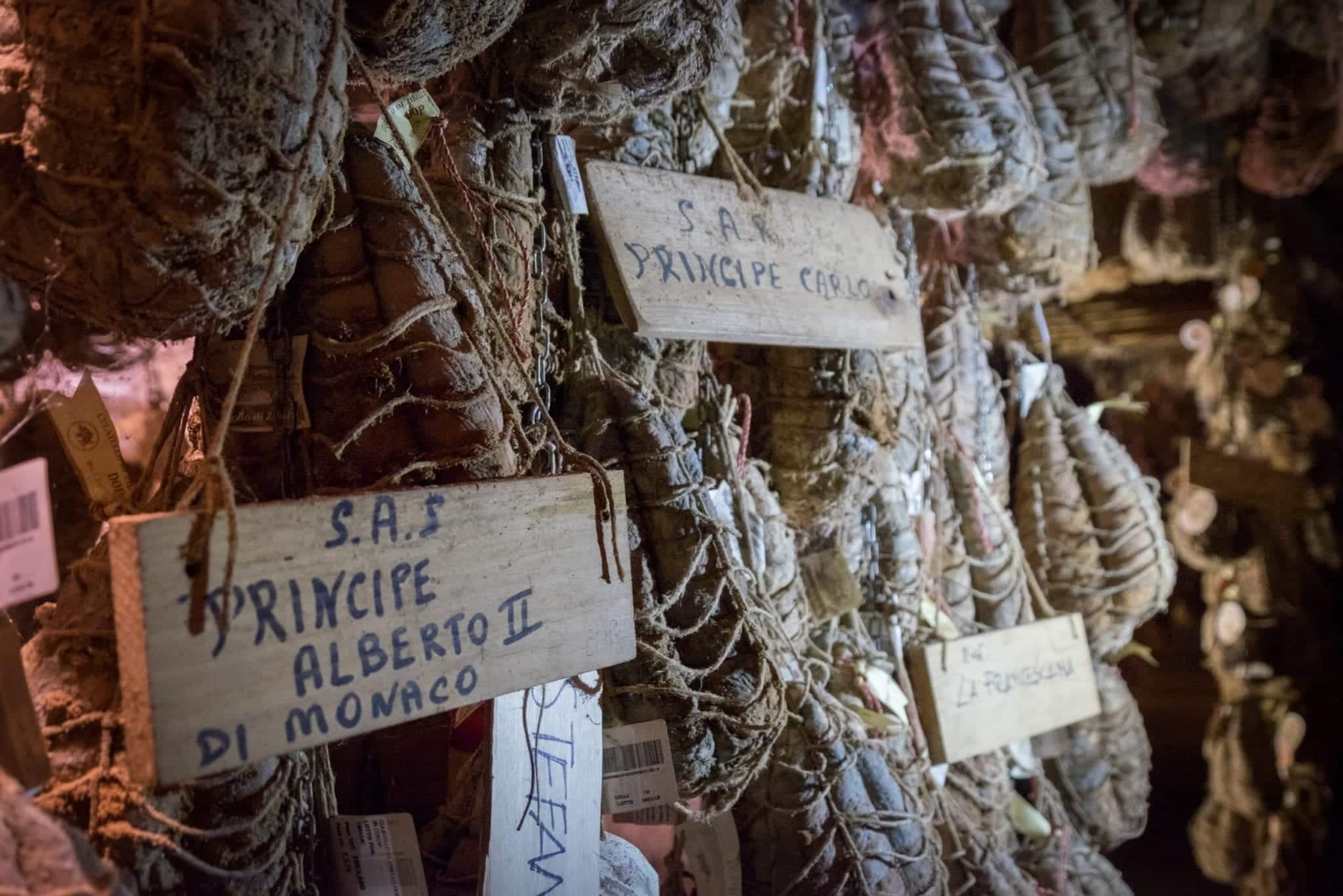 Hanging culatello in the basement, some labeled with "Principe Alberto II" and "Principe Carlo" for Prince Albert and Prince Charles.