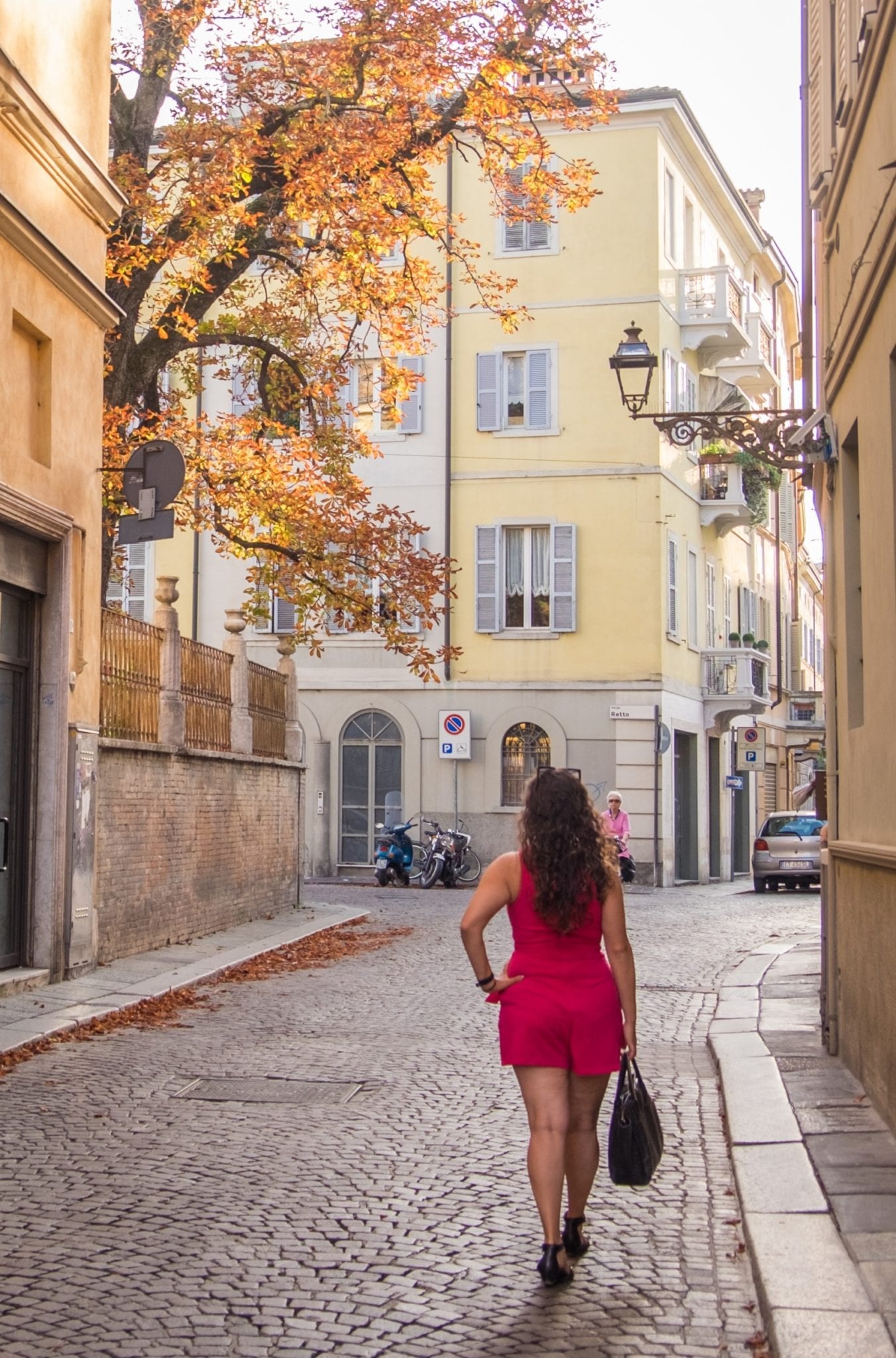 Kate wears a short hot pink romper and stands on a cobblestoned street in front of a yellow building and a tree with autumn leaves.