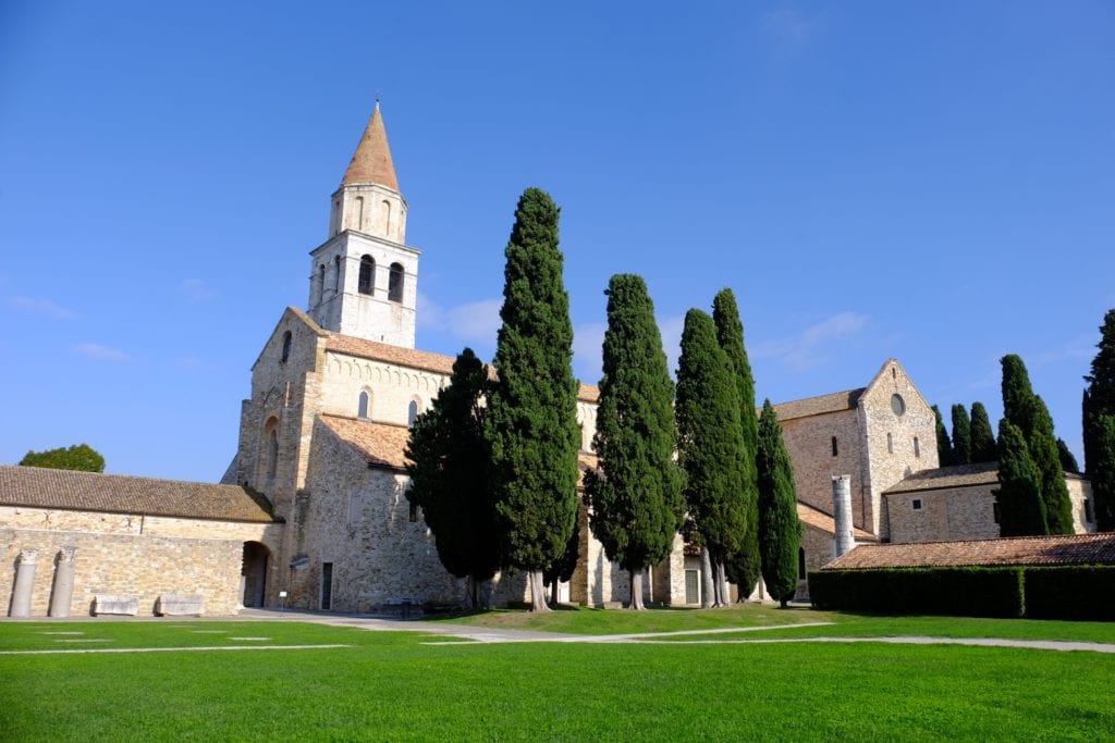 The cathedral in Aquileia, Italy, a small church with an orange roof with tall cypress trees in front.