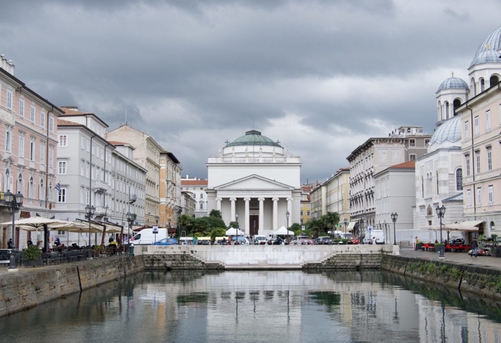 A white building with columns behind a canal, underneath a cloudy gray sky in Trieste, Italy.
