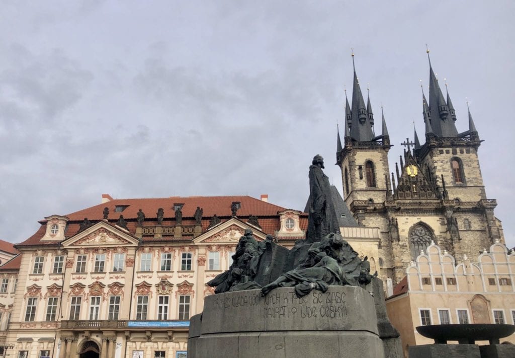 A statue in front of a church tower and crenellated building on Old Town Square in Prague.