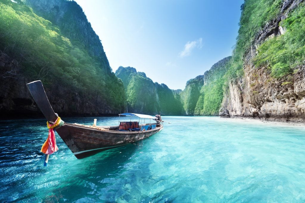 A longtail boat cruising through clear turquoise water surrounded by bright green cliffs.