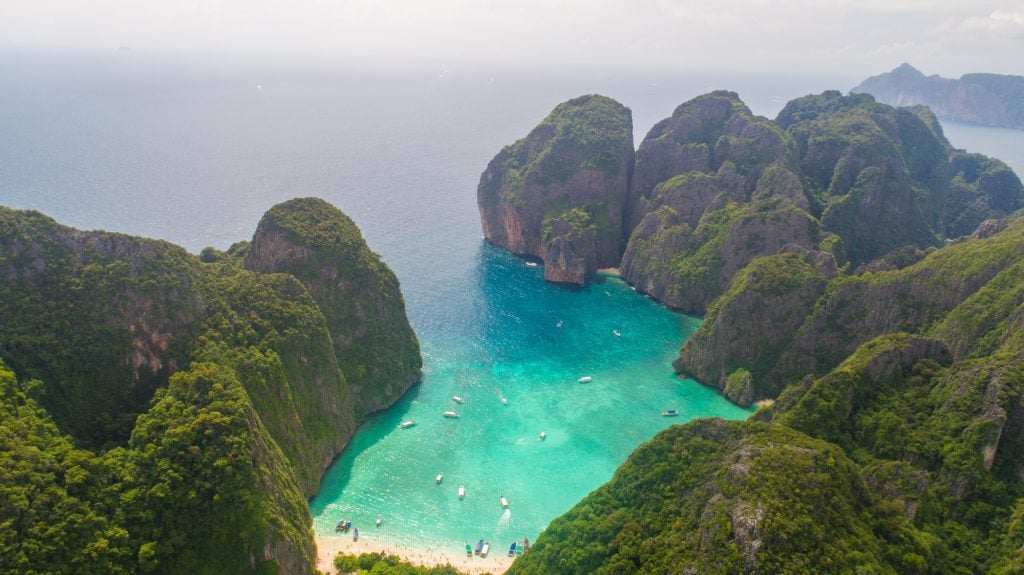 An aerial view of a turquoise bay and secluded beach in Thailand surrounded by green mountains.