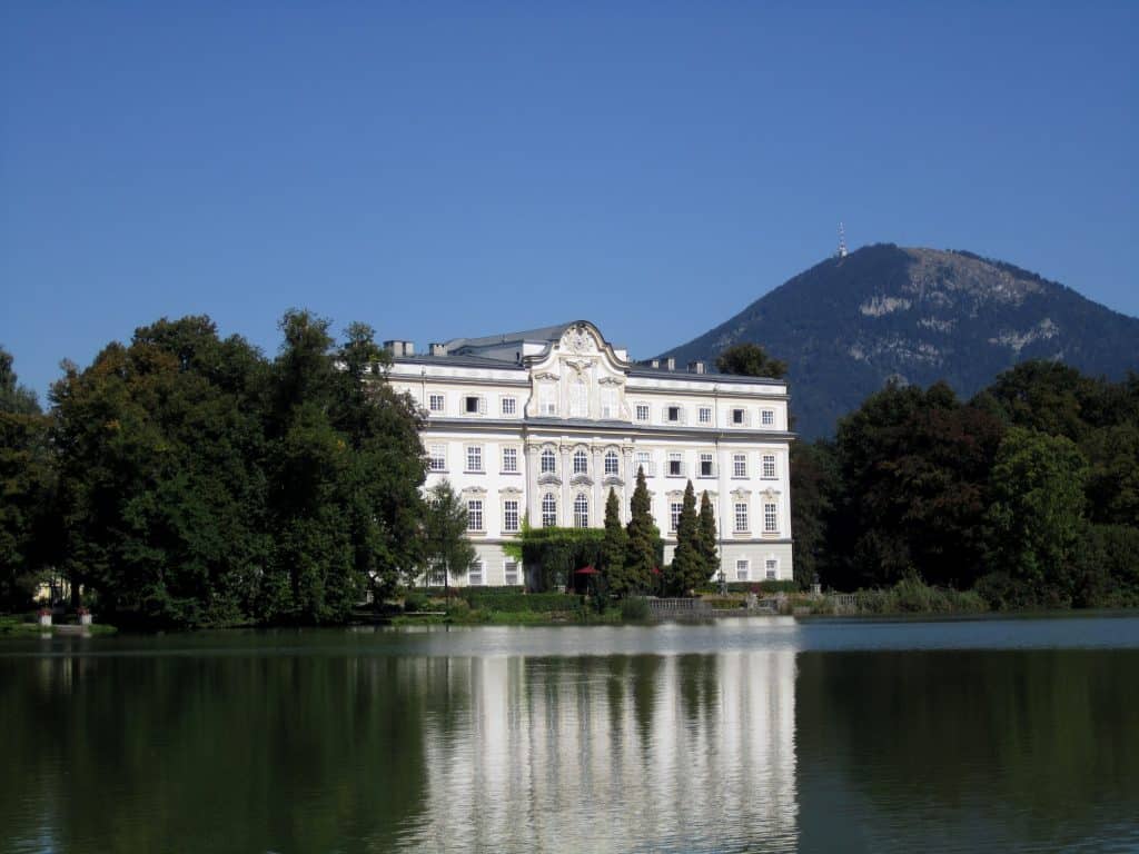 A big white mansion perched on a still lake, reflecting in the water.
