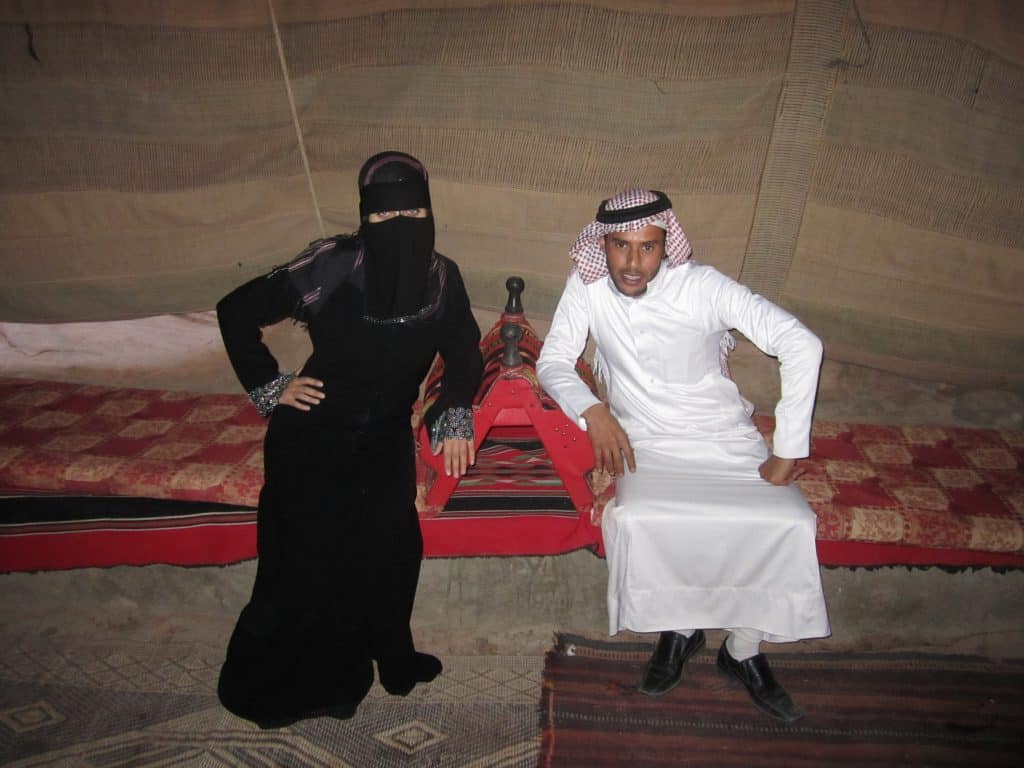 Kate in a black Bedouin gown with a black mask hiding most of her face, sitting next to Hasan in a long white garment and a red and white scarf hanging on his head.