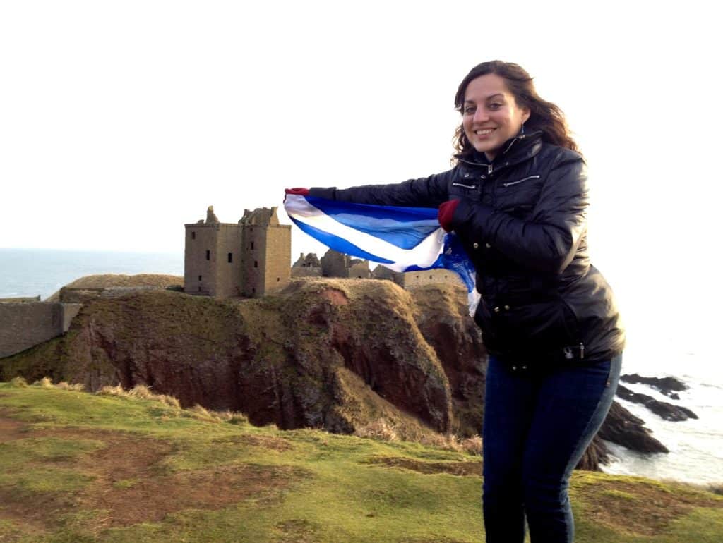 Kate holding a blue and white Scottish flag in front of a old stone castle on a cliff.