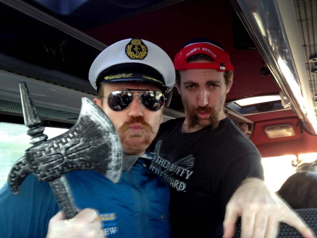 Two guys on a bus, rocking handlebar mustaches, one in a captain's hat and holding a fake axe.