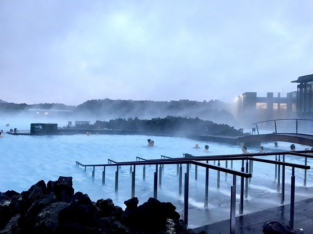 People swimming in Iceland's Blue Lagoon at dusk, steam rising up from the milky blue waters.