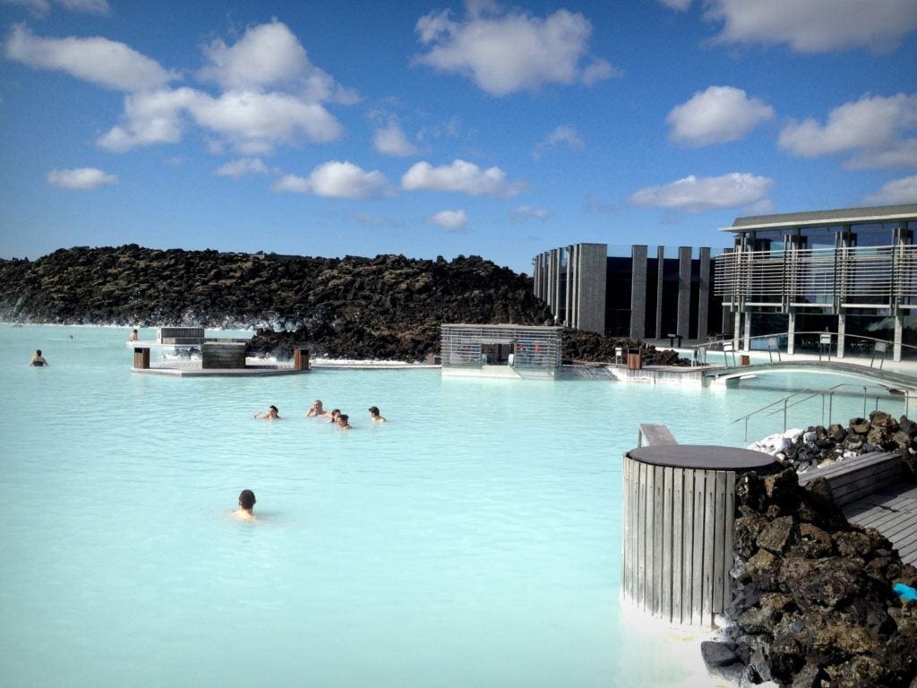 The pale blue water of the Blue Lagoon, a few people swimming in it, underneath a white and blue sky.
