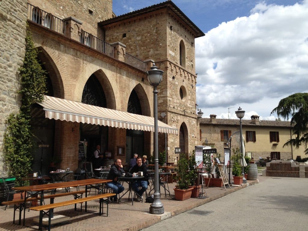 Men sitting at an outdoor sidewalk cafe in Italy, in front of a medieval-looking building.