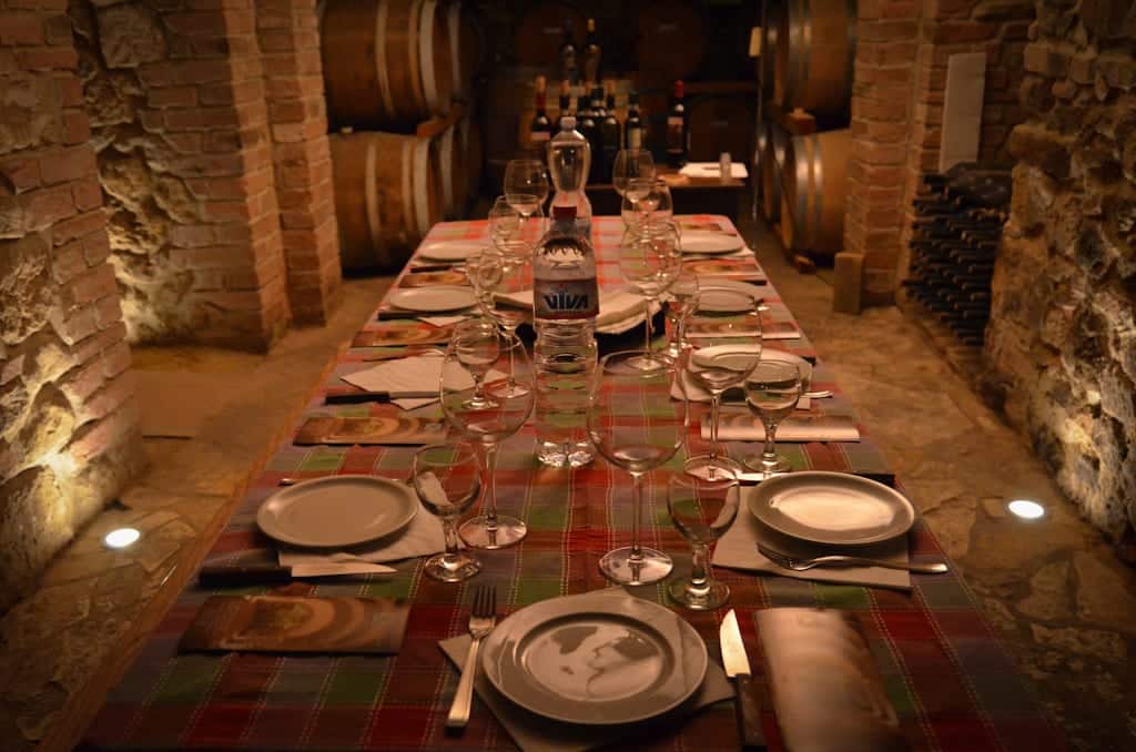 A big table set in a wine cellar with lots of wine glasses for each person.