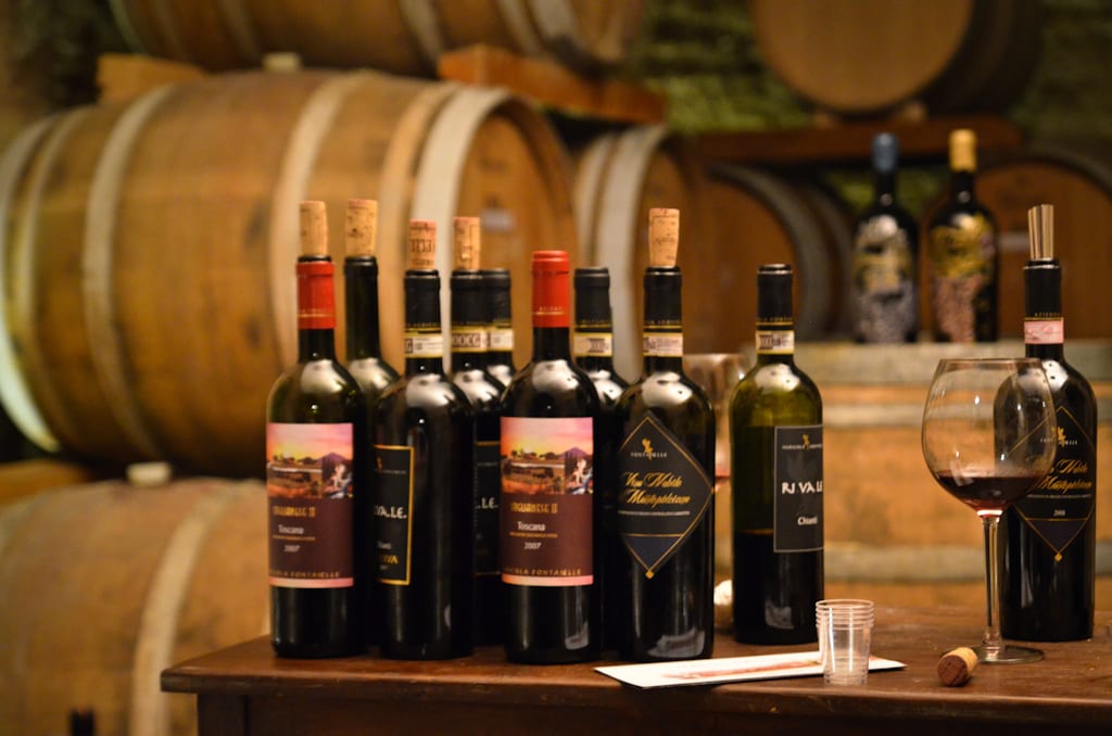 Several wine bottles lines up on top of a wine barrel, more wine barrels in the background.