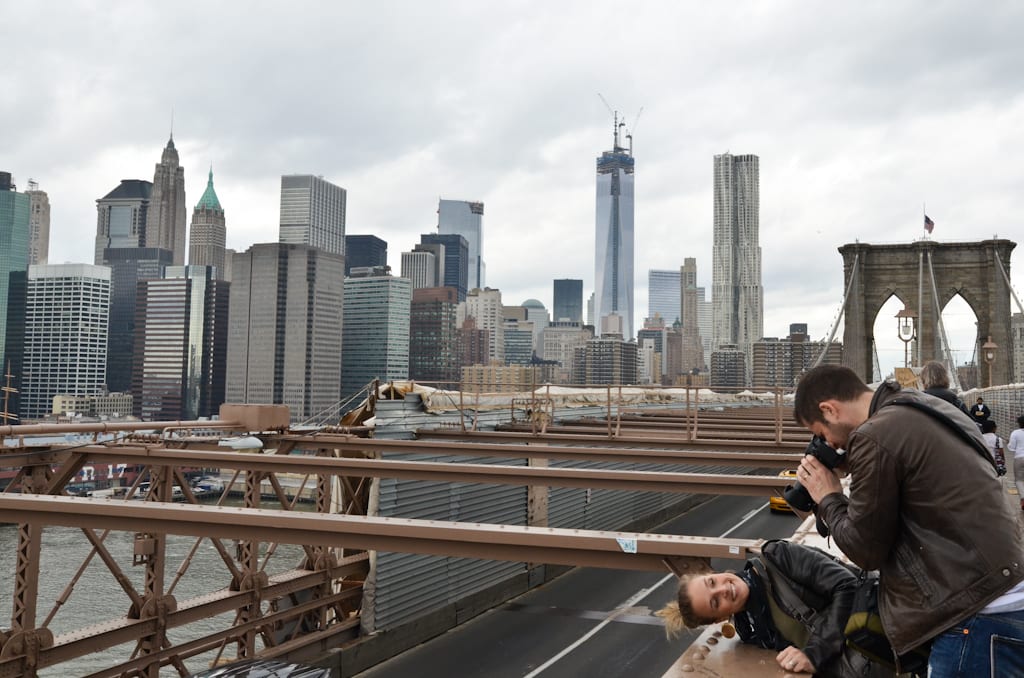 A couple leaning over the edge of the Brooklyn Bridge to take a picture, the Manhattan skyline behind them.