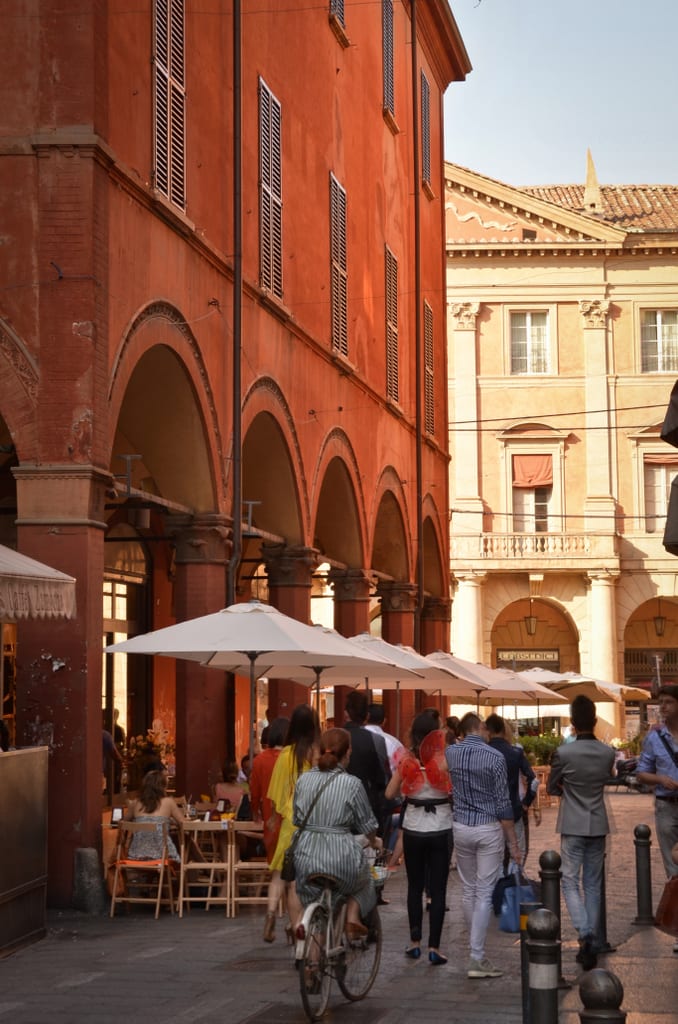 A group of people walking past a sidewalk cafe in Bologna next to a bright red building with porticoes.