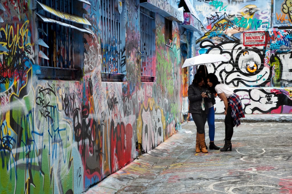 An alley filled with graffiti, with women under an umbrella looking at their phones.