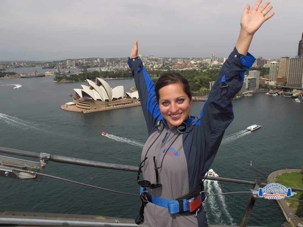 Kate smiling with her arms up, wearing a jumpsuit and standing on the Sydney Harbour Bridge, the Opera House on the ground in the background.