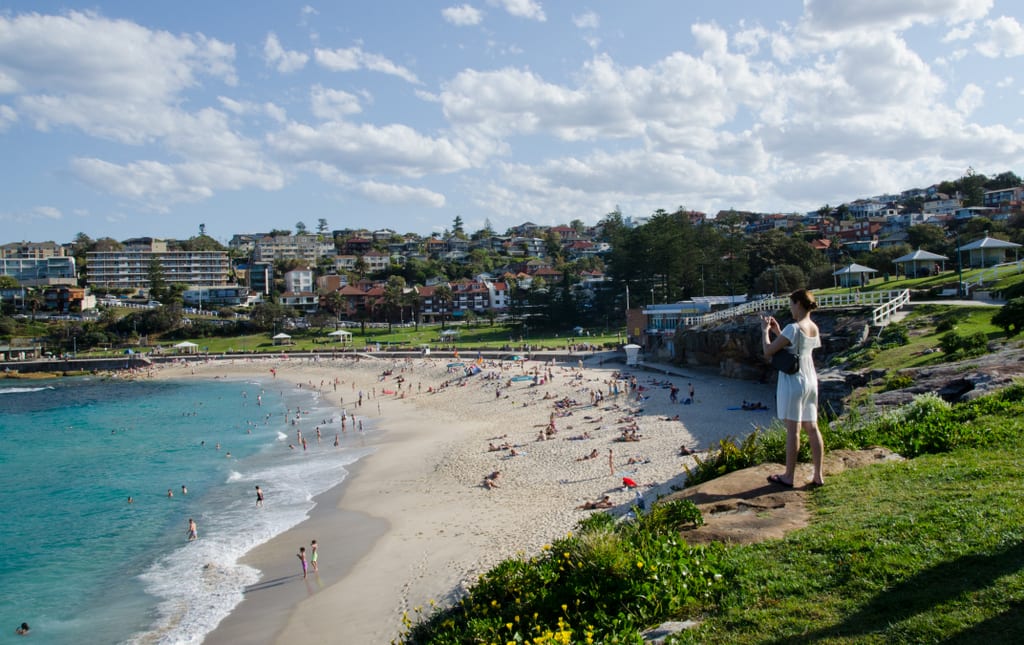 A woman in a white dress standing on a cliff and taking a picture of the beach in Bronte, Sydney. The water is bright turquoise and the sky is pale blue streaked with clouds.