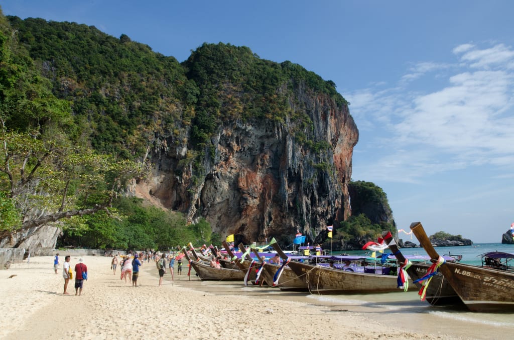 Railay Beach, people walking in the distance with long tail boats on shore and cliffs the distance.