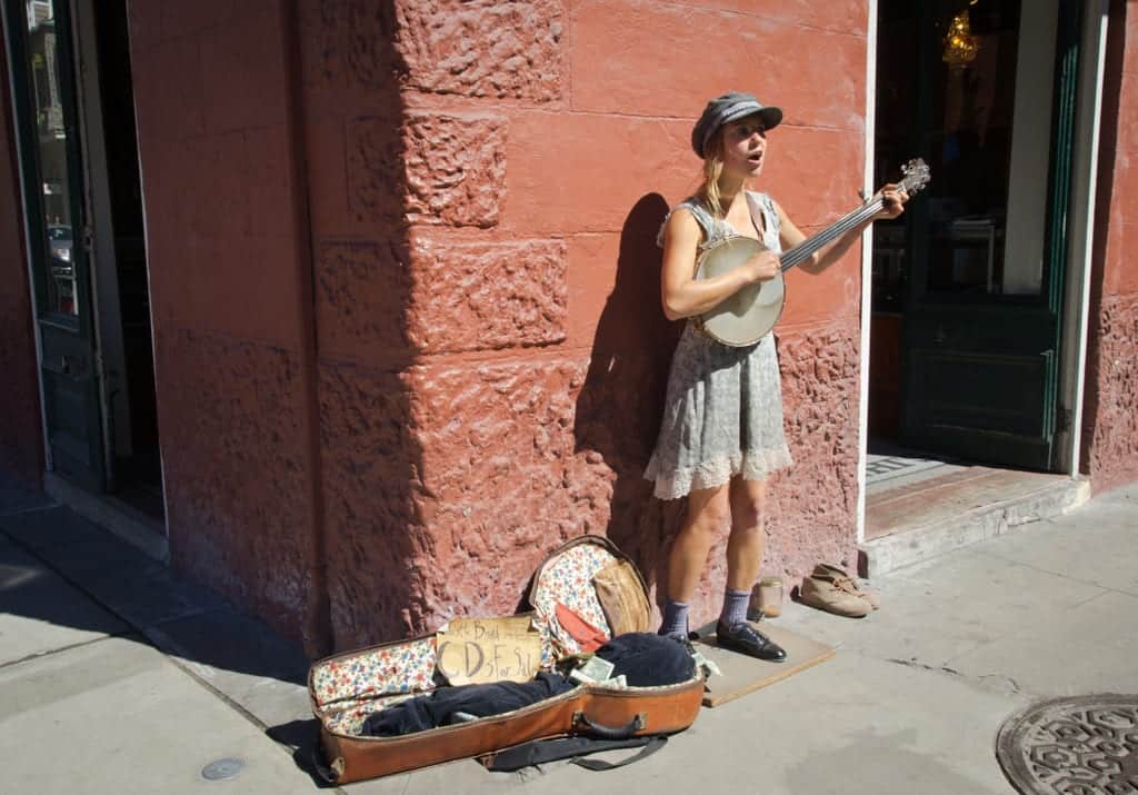 A woman wearing a dress and playing a small guitar on a street in New Orleans, an open case on the ground next to her for tips.