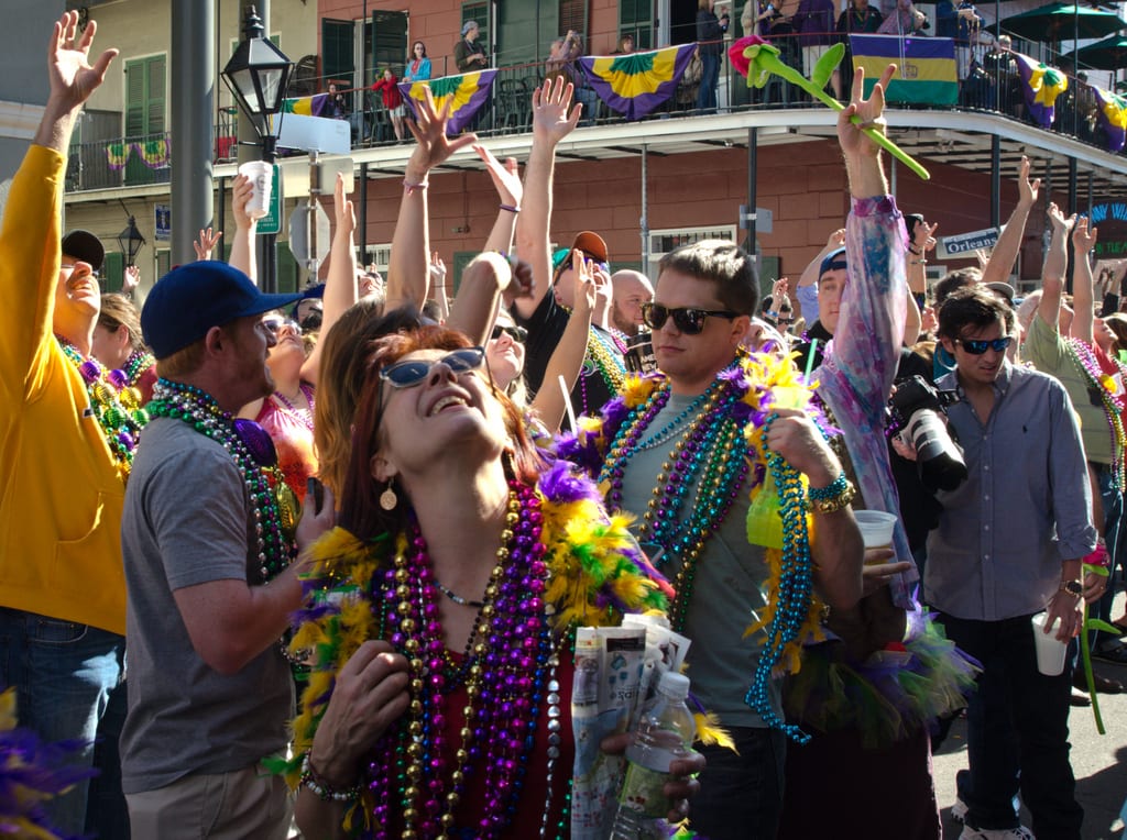 A crowd in New Orleans reaching up to a balcony to catch beads. A woman in sunglasses looks up at the balcony and smiles, lost in the moment.