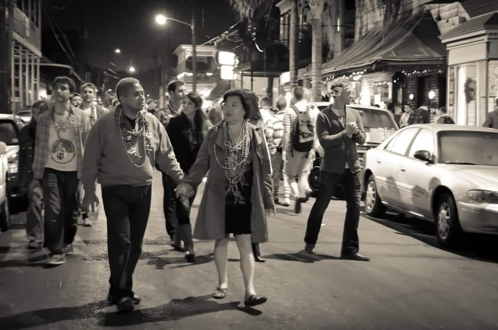 A black and white shot of a couple walking hand in hand, mardi gras beads around their neck, down Frenchman Street in New Orleans, where crowds are waiting to get into music bars.