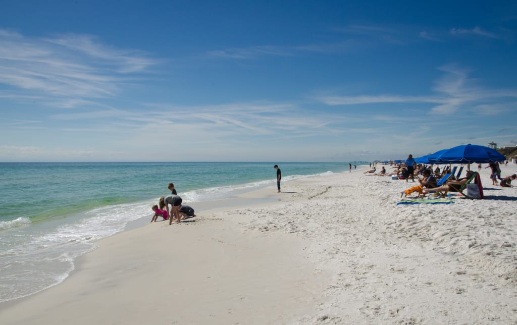 People sitting on the white sand beach, some kids playing near the water, other adults sitting in sun loungers underneath umbrellas.