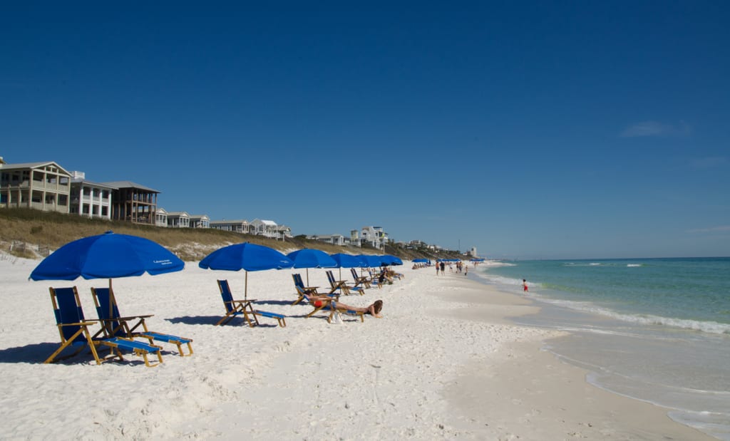 People lying in beach chairs on the beach in Seaside, Florida, underneath blue umbrellas.