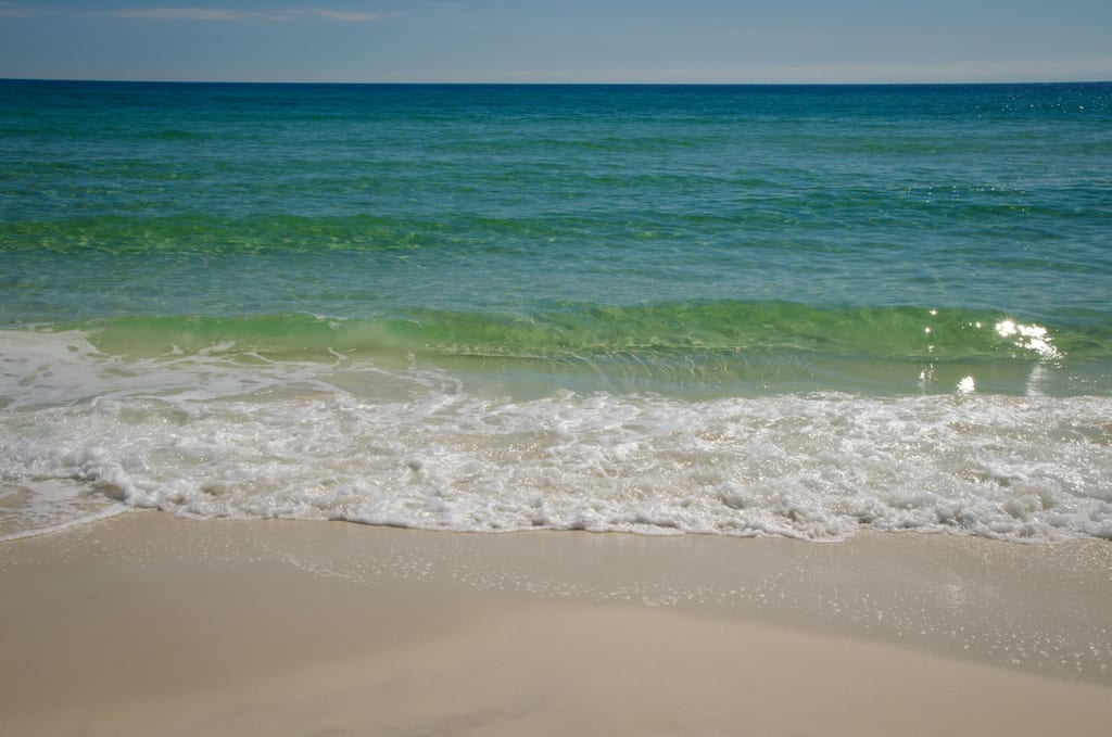 Bright blue-green clear ocean water, foamy white waves crashing on the light brown sand.