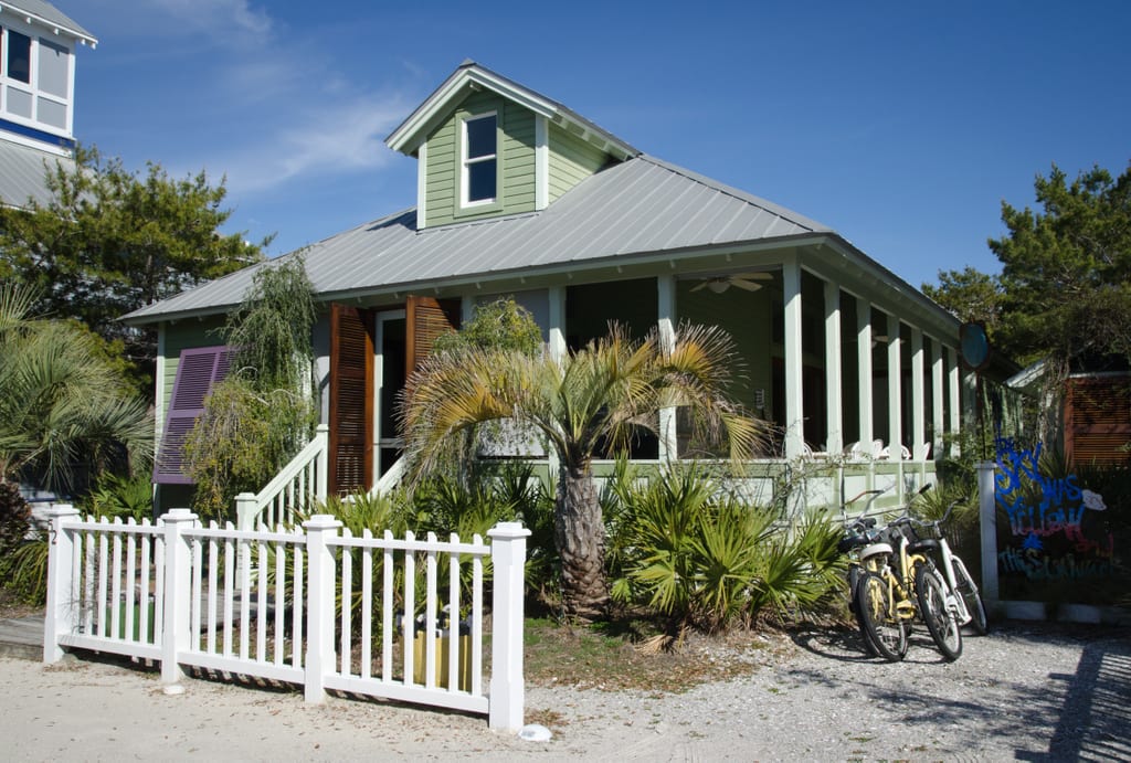 A green bungalow surrounded by small palm trees with a white picket fence.