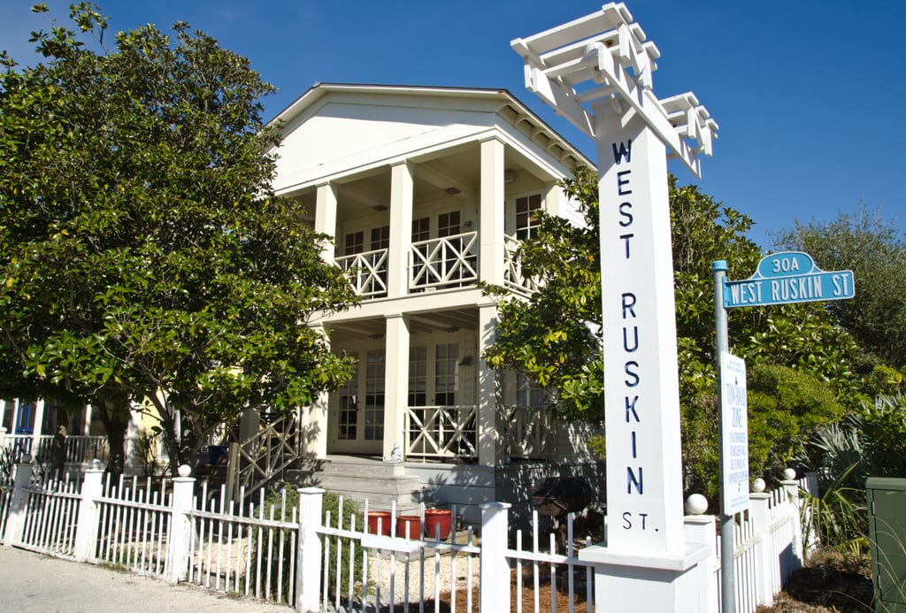 A tall beige house with white balconies and columns. There is a white fence. It's next to a sign reading West Ruskin St.