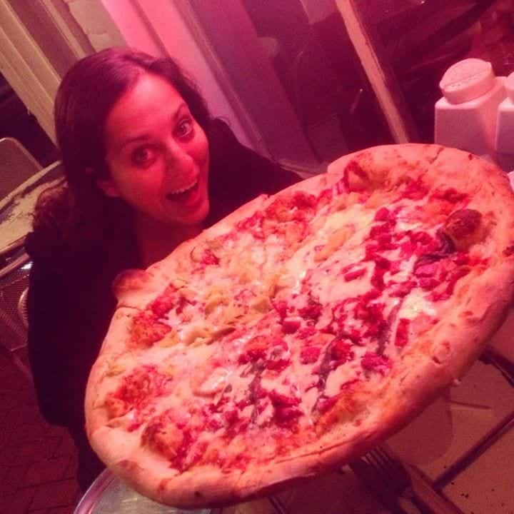 Kate about to take a bite of an ENORMOUS round pizza, fear and excitement in her eyes.