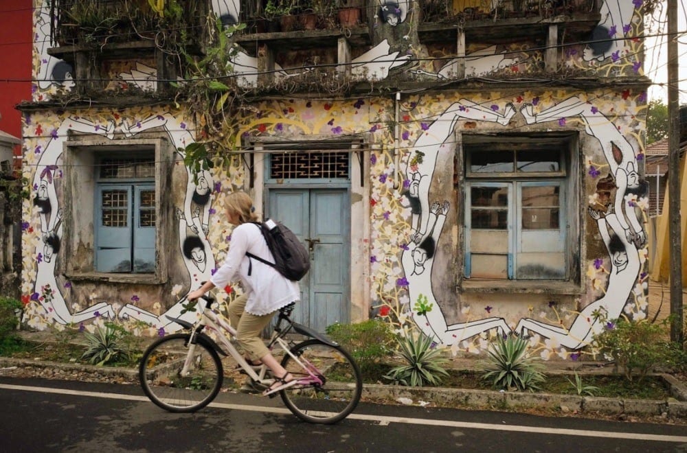 Blonde woman riding a bicycle against a painted wall with a blue door in Kochi, India.