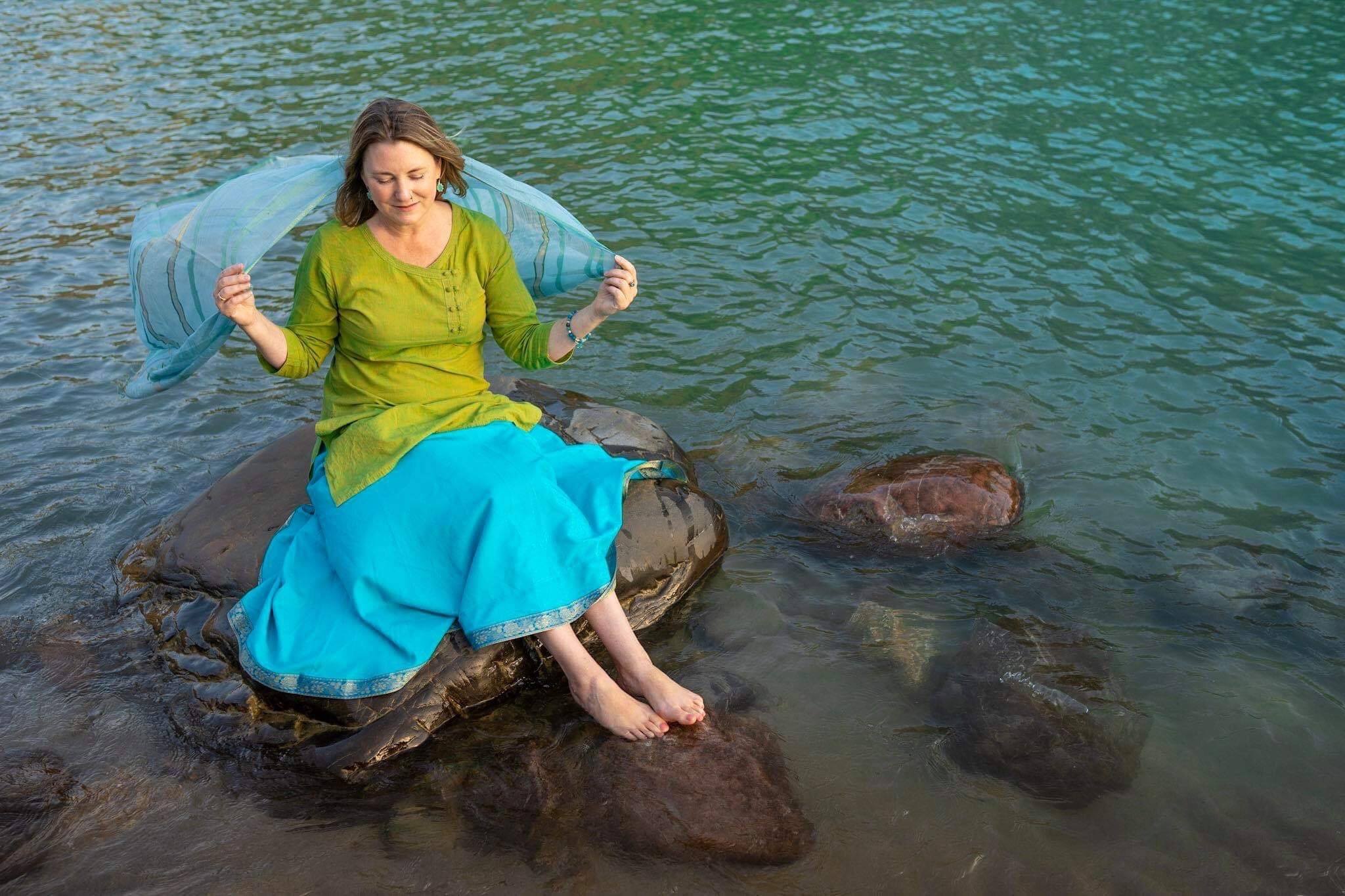 Mariellen Ward in a green and blue dress holding a blue shawl behind her, sitting on a rock in the Ganga river.