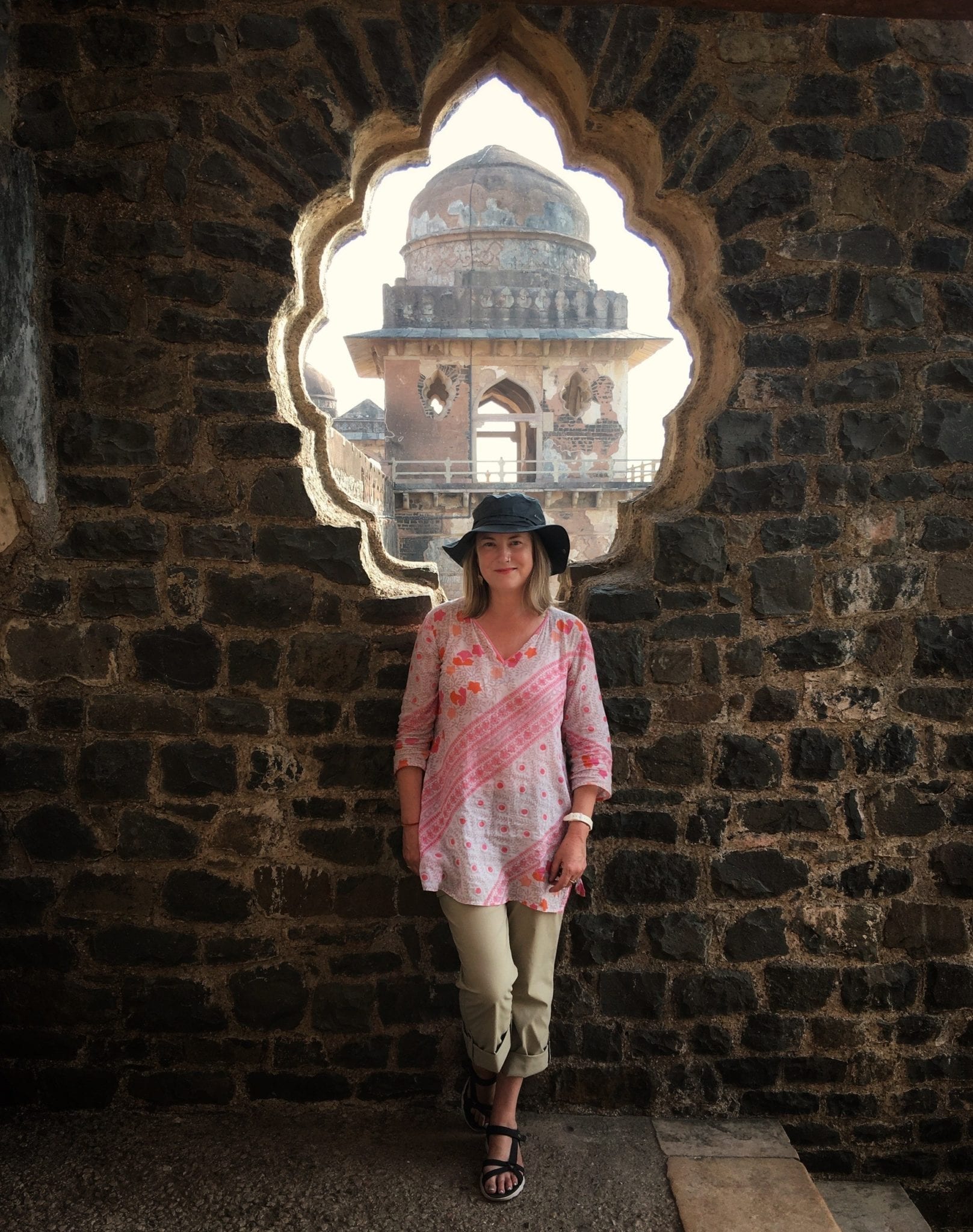 Mariellen Ward wearing a white and red top and black hat and posing in front of a scalloped window opening in a temple in Mind, India. 