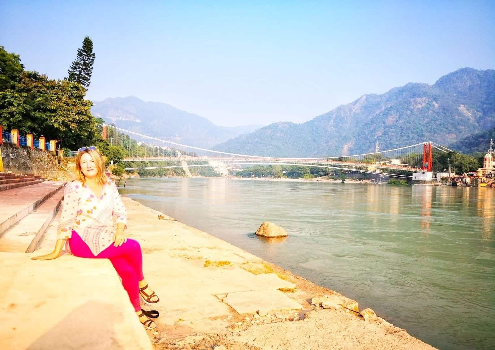 Mariellen Ward wears a white Indian top and pink trousers and poses on the banks of the Ganga river in Rishikesh, mountains behind her.