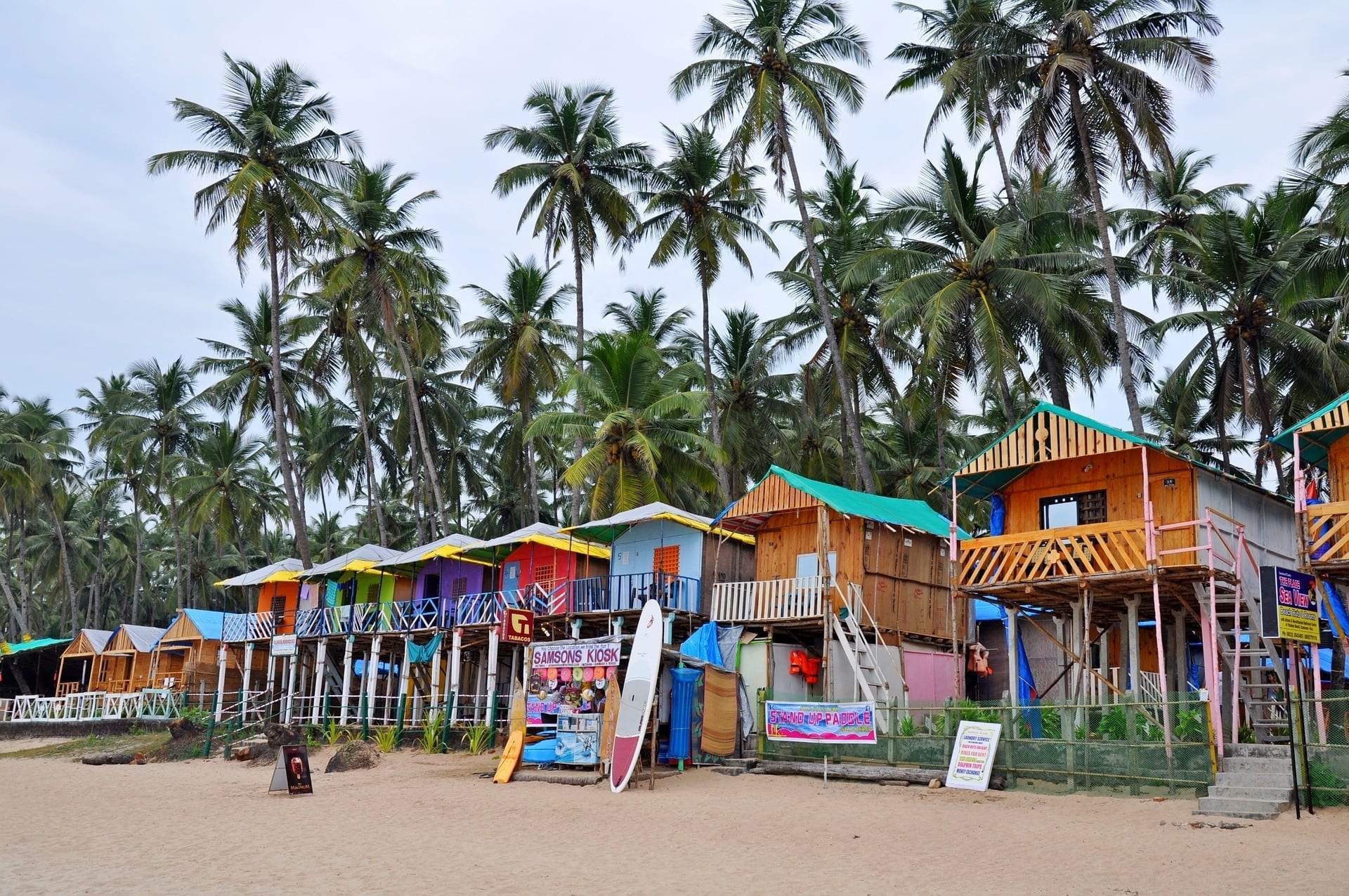 A line of brightly painted beach shacks beneath palm trees on the sand in Goa, India.