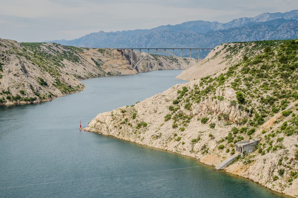 A river in Croatia, with gray-green steep hills rising up on each side. You see a steel suspension bridge across the river in the distance. Cloudy sky and mountains in the distance.