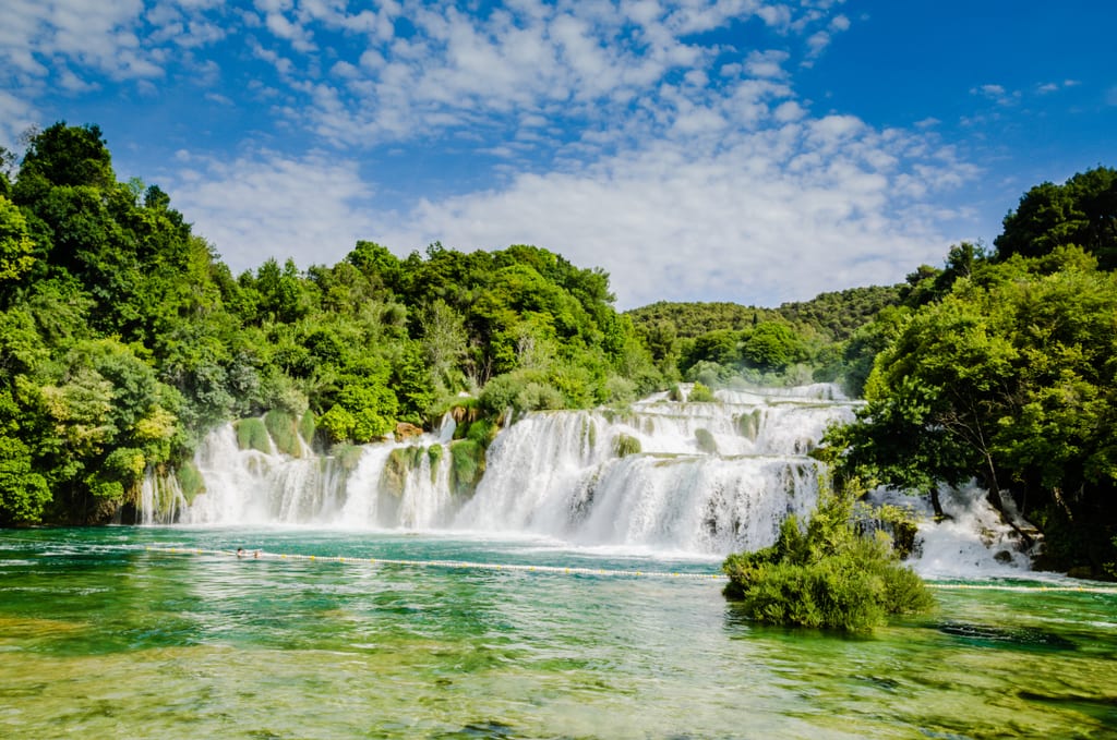 A long white waterfall with several streams, flowing into a green lake, in the middle of a forest, underneath a blue and white cloud streaked sky.