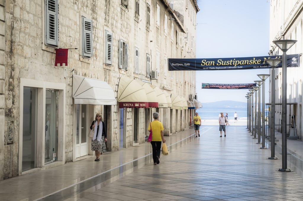 A quiet street in Split early in the morning surrounded by pale gray stone buildings, the ground so smooth it's reflective, the sea in the distance.