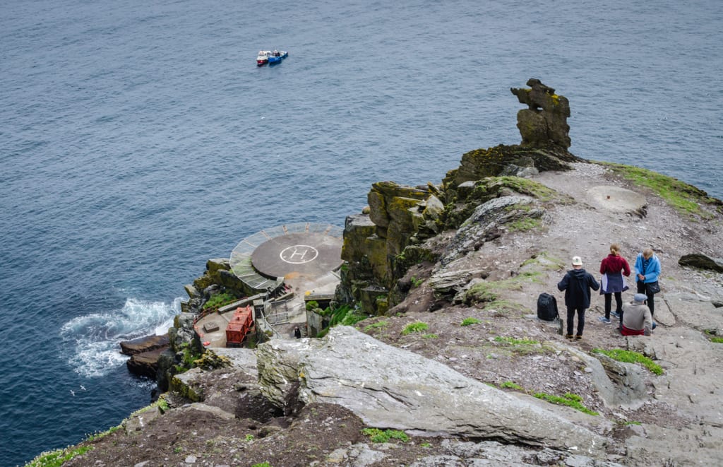 Looking down toward a helipad with an H in the middle of it at the bottom of Skellig Michael near the ocean. Further up is a rocky landing, and you see four people pausing before climbing the stairs.