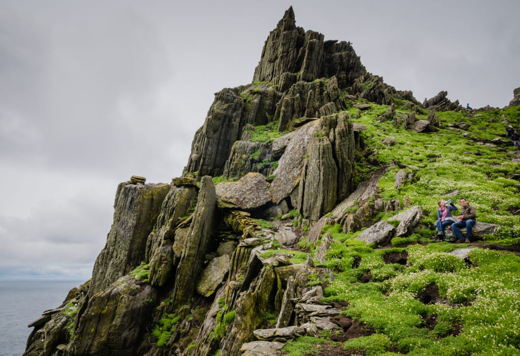 The top of Skellig Michael: jagged pointy rocks pointing toward the gray cloudy sky. The ground is covered with moss and tiny white flowers. A couple sit on a slab of rock and drink form their water bottles.