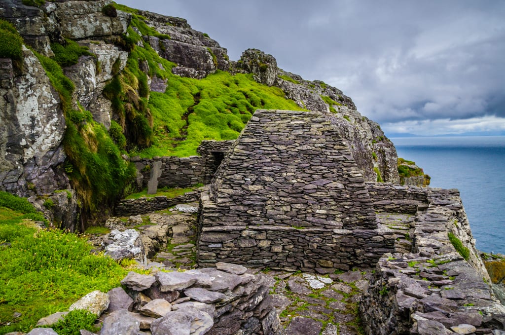 A beehive-shaped rocky hut in front of a steep green mossy hill.